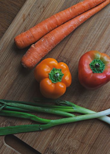 A vertical shot of fresh carrots, bell peppers, and spring onions on a wooden board