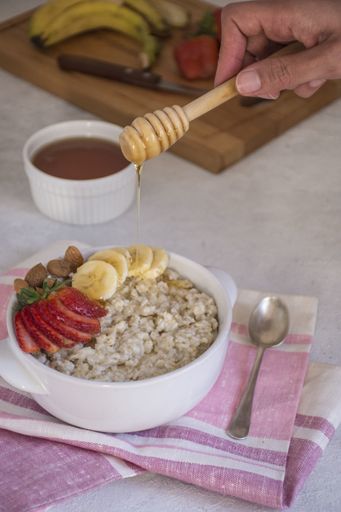 A vertical high angle shot of a person pouring honey on a bowl of oatmeal with fruits