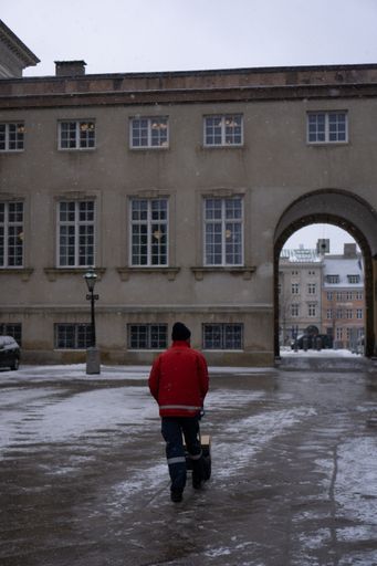 A back shot of a man with a red coat walking on a snowy ground near a building in Copenhagen, Denmark