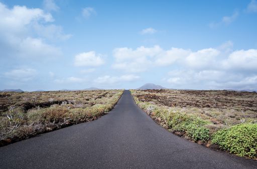 A scenic view of an empty asphalt road surrounded by a dried field under a blue cloudy sky in Lanzarote