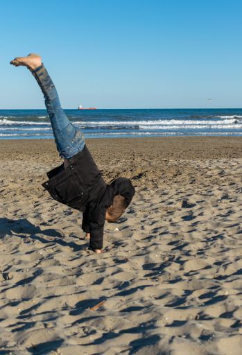 A vertical shot of a Caucasian boy doing parkour on the sandy beach against ocean waves