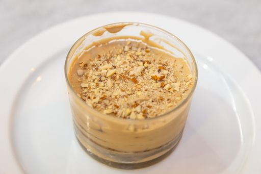 A close-up shot of a pudding dessert with pieces of walnuts on top, in a glass cup, on a white plate