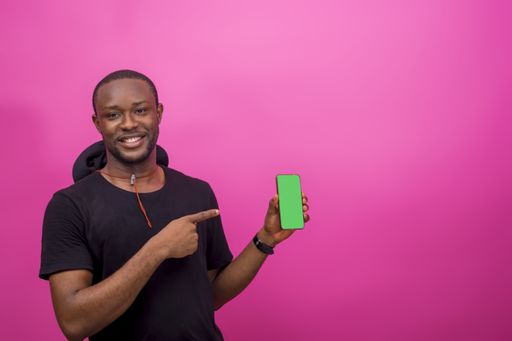 A young African male holding and pointing a cellular phone isolated on a light pink background