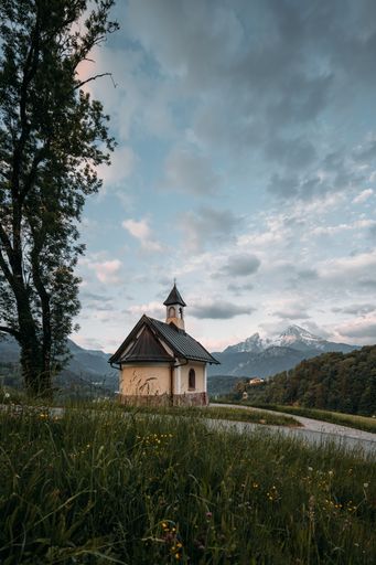 A view of a small church in the meadow or park surrounded by trees and mountains in the distance