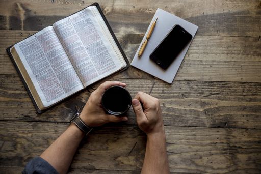 An overhead shot of a male holding his coffee mug near a book and smartphone on a wooden surface