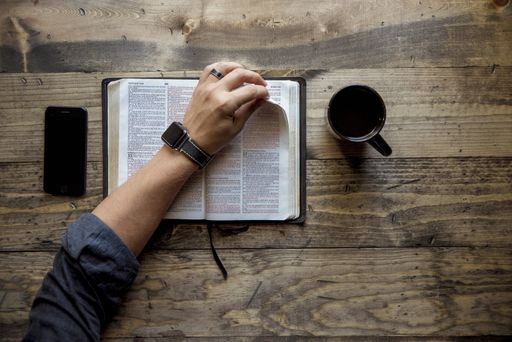 An overhead shot of a person flipping the page of a book near the coffee and a smartphone on a wooden table