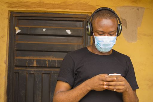 A young African-American male wearing a mask and a headset while using a phone in-front of the door
