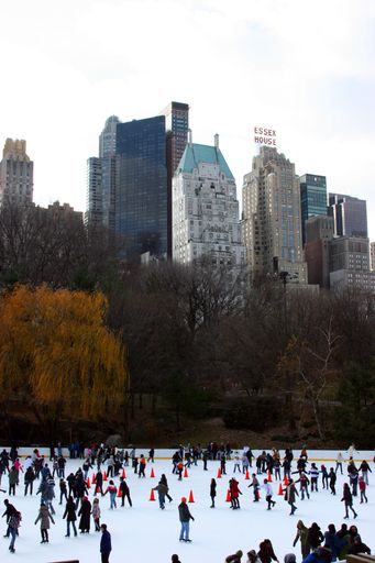 A vertical shot of people ice skating with NYC buildings in the background