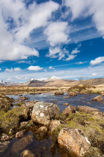 A vertical shot of a river with tall mountains and a cloudy blue sky in the background