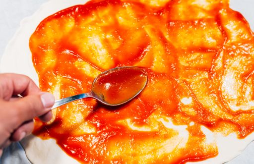 A high angle shot of a person adding sauce on pizza dough with a spoon in the kitchen