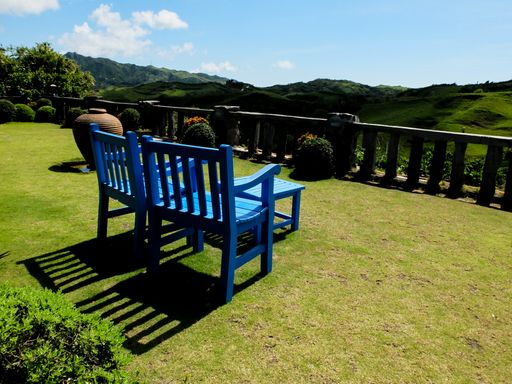 A shot of two blue wooden chairs and a small table in the park with a beautiful view of the green mountains against blue sky in bright sunlight
