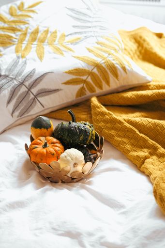 A vertical shot of small pumpkins in the bowl near the orange blanket on the white sheet