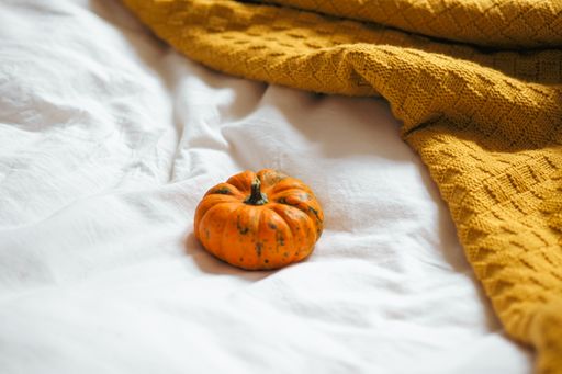 A closeup of a single orange pumpkin on a bed- white bedsheets, yellow ochre knitted blanket