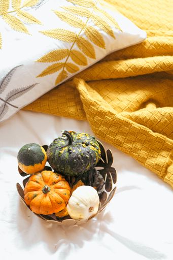 A vertical high angle shot of a variety of pumpkins in s wooden bowl on white bedding with an ochre blanket and an embroidered pillow