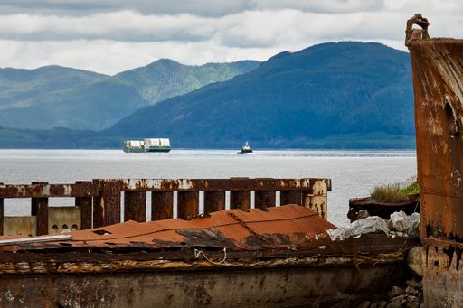 Looking over old rusty ships that form a breakwater to a tug pulling a barge loaded with containers down the strait with mountains in the background.
