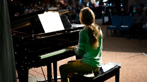 A little girl playing the piano in front of an audience captured from the back