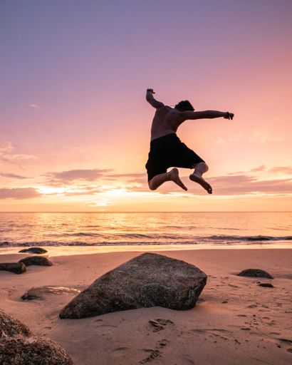 A back view of a man jumping high on the sandy beach on the coast