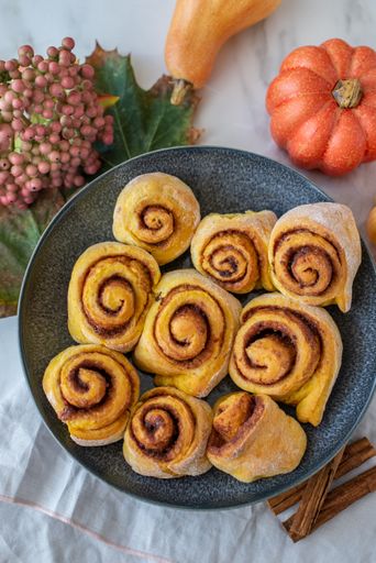 A top view of cinnamon rolls on a black plate with cinnamon sticks and pumpkins next to