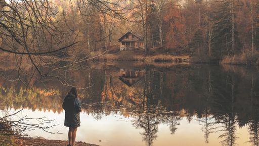 A lonely woman standing near the lake with the reflection of the isolated wooden cabin and trees visible