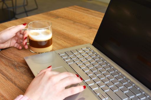 A closeup of a woman's hand working on a laptop and holding a cup of cappuccino on the other hand