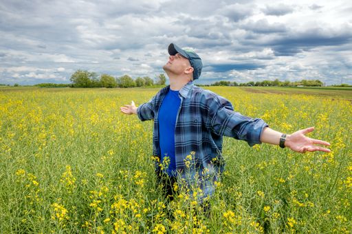 Farmer standing in a rapeseed (oilseed) field, spreading his arms and looking at the sky with heavy rain clouds above his head.