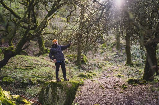 A young male stanging on a big rock and taking a selfie in a lush forest during the hike