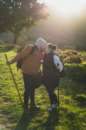 A vertical shot of a couple kissing and taking pictures on a green mountain path during the hike on a sunny day