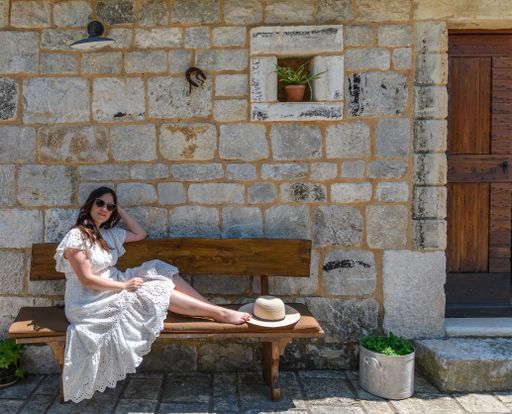 A young Caucasian woman wearing a white dress, sitting on a bench in front of an old stone house