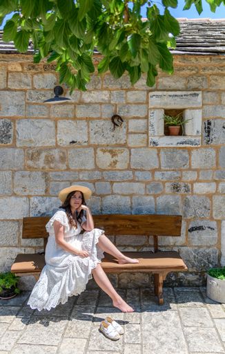 A young Caucasian woman wearing a white dress, sitting on a bench in front of an old stone house