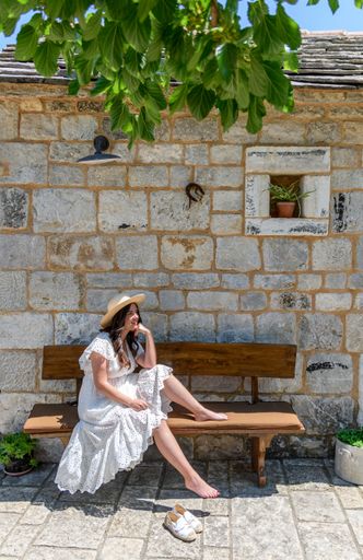 A young Caucasian woman wearing a white dress, sitting on a bench in front of an old stone house