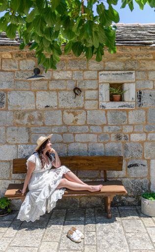 A young Caucasian woman wearing a white dress, sitting on a bench in front of an old stone house