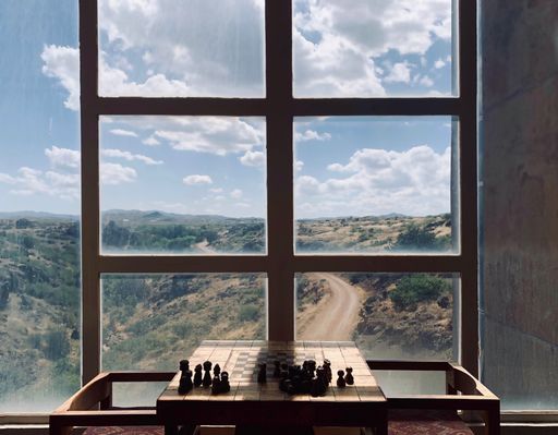 A chessboard with black pieces behind a window with the view of a mountain road surrounded by bushes on a blue sky background