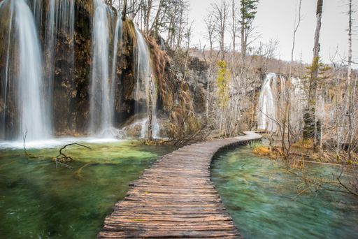 A horizontal shot of a wooden pathway on a body of water with waterfalls flowing from the cliffs