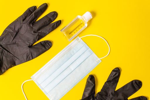 An overhead shot of a medical mask, black medical gloves, and a sanitizer on a yellow background