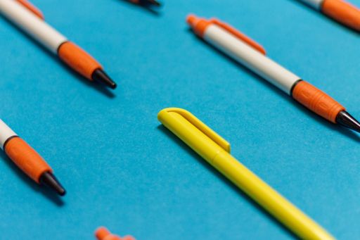 A closeup shot of a yellow pen in identical orange and white pens on a blue background