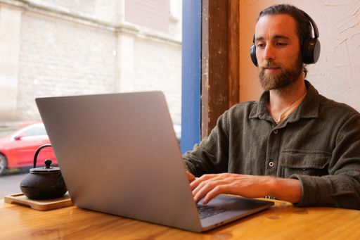 A man with beard working with computer and headphones sit near the window
