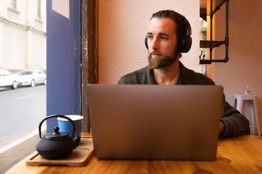 A man with beard working with computer and headphones sit near the window