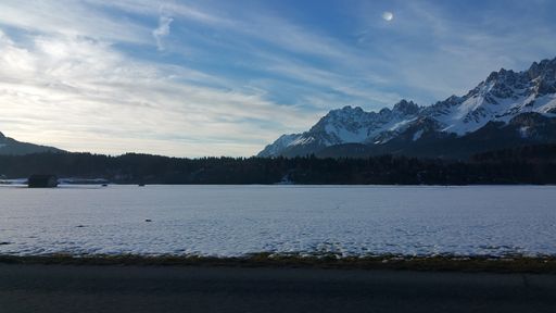 Snow-covered alps mountains in a sunny evening winter day. Clouds and moon. POV first person view shot on a asphalted mountain road.