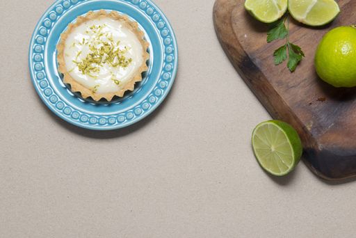 A top view of a key lime tartlet on a plate and key lime fruits on a wooden board