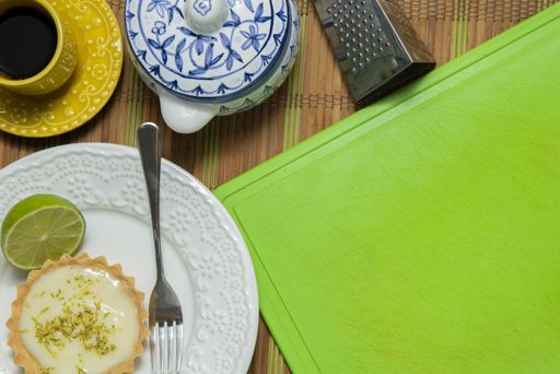 A top view of a key lime tartlet and a fork on a plate with tea set, grater and green slicing board