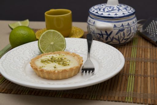 A soft focus of a key lime tartlet and a fork on a plate with key lime fruits, tea set and grater in the background on a bamboo mat