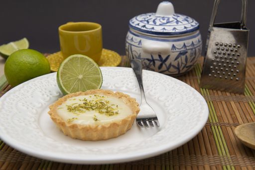 A soft focus of a key lime tartlet and a fork on a plate with key lime fruits, tea set and grater in the background on a bamboo mat