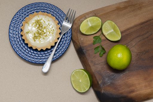 A top view of a key lime tartlet and a fork on a blue plate and key lime fruits on a wooden board