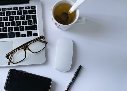 A high angle shot of a laptop, glasses, a cellphone and a cup of tea on a white surface