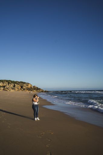 A vertical shot of a female standing on the beach while taking a photo of the beautiful ocean
