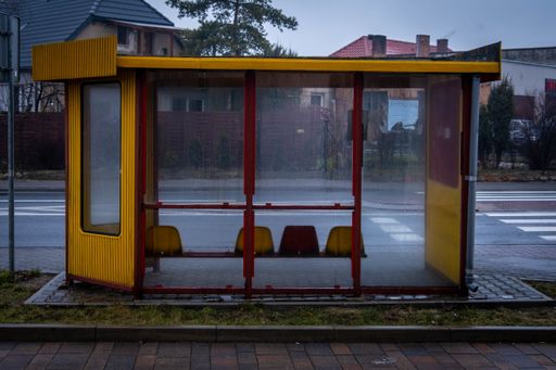 A yellow and red bus stop on the sidewalk in an urban area with a missing seat