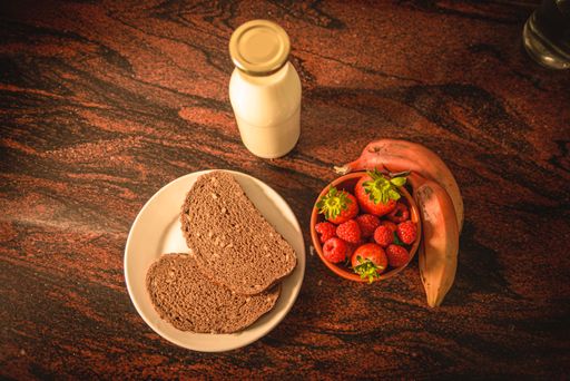 A top view of a bowl with berries, two pieces of rye bread, banana and a bottle of milk on the table
