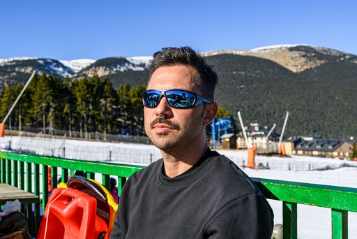 A young caucasian male with facial hair and sunglasses sitting under the sun in a snowy park