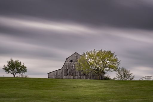A dramatic shot of an old wooden barn standing in a field next to trees under a dark cloudy sky