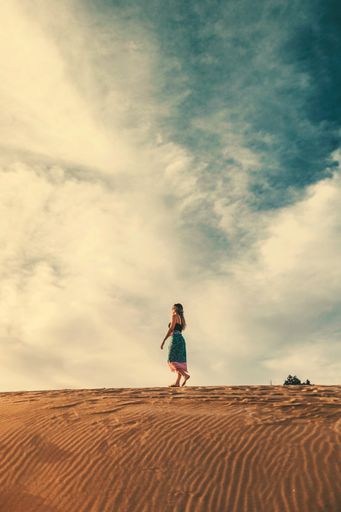 A vertical shot of a woman in a dress walking on sand barefoot during daytime